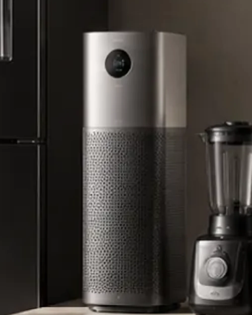 A cylindrical tower air purifier in brushed silver and dark mesh, standing on a concrete plinth.