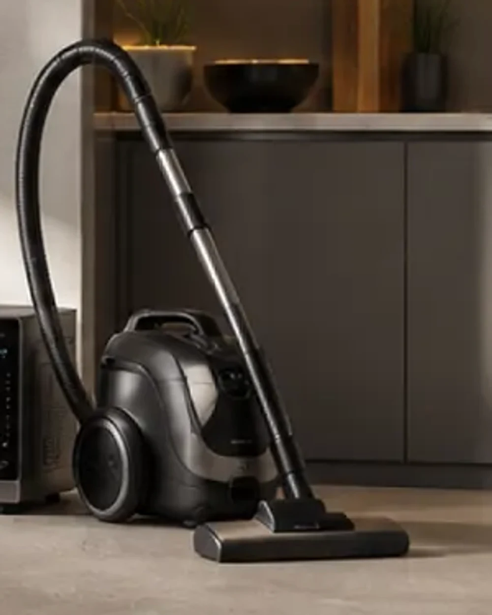 A dark grey cylinder vacuum cleaner with a chrome telescopic wand and a wide floor head.